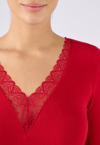 Top rosso a maniche lunghe con scollatura a V, caratterizzato da un intricata rifinitura in pizzo, texture liscia e design aderente. Il collo mette in risalto i dettagli dei motivi in pizzo.