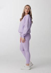 Fialová mikina s elastickými manžetami a logem, spárovaná se shodnými žebrovanými legínami. Bílé tenisky dotvářejí ležérní outfit.