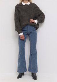 Jersey amplio de punto en gris oscuro con una camisa blanca de cuello debajo, combinado con pantalones acampanados azul claro y tacones negros puntiagudos.