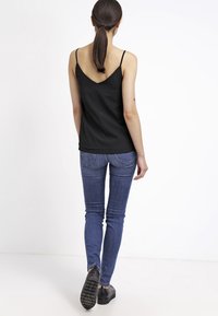 Top nero senza maniche con schiena a V, abbinato a jeans blu attillati e scarpe nere. L'outfit presenta una texture di tessuto liscia.