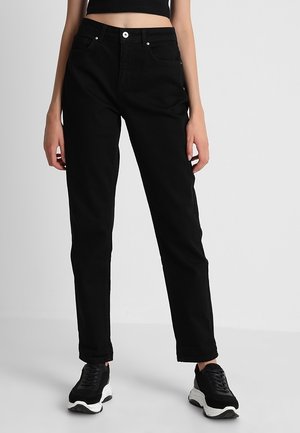 Person wearing high-waist black jeans and black sneakers with white soles, standing against a plain light gray background.