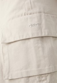 Beige szövet cargo nadrág nagy zsebbel, textúrált pamut felület látható. A "OBEY" szó fehér hímzéssel van a zseb fedelén.