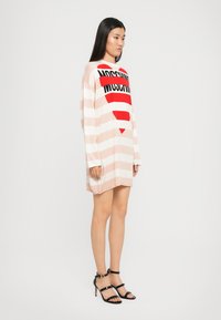 Abito lavorato a maglia a righe rosa e bianche con un grande cuore rosso e il testo "Moschino" in nero. Maniche lunghe, vestibilità ampia e orlo leggermente curvato.