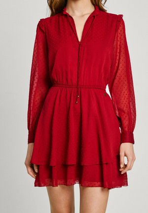 Vestido rojo hecho de tela transparente con textura de puntos, cuello con solapa, mangas largas y dobladillo con capas de volantes. Detalle de cintura con cordón.