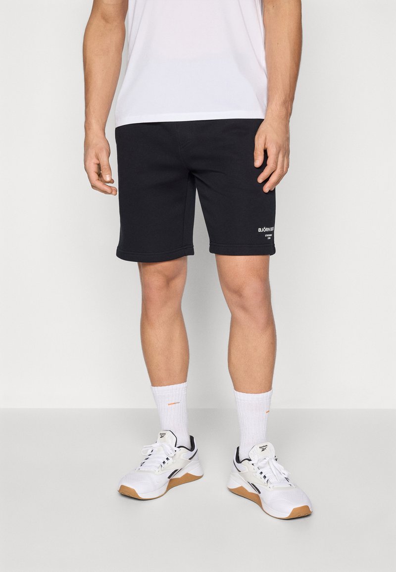 Črne športne kratke hlače z ohlapnim krojem, rebrastim pasom in belim logotipom. Obute imajo bele teniske in nogavice do gležnjev.