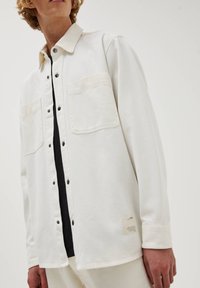 Biała koszula zapinana na guziki z dwoma kieszeniami na piersi, czarnymi guzikami i miękką teksturą. Posiada metkę na bocznej szwie, noszona na czarnej bluzce.