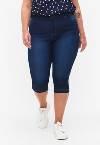Pantalones capri de mezclilla azul oscuro con una cintura de media altura, presentando un diseño ajustado y detalles cosidos. Combinados con zapatillas blancas y una blusa de lunares.