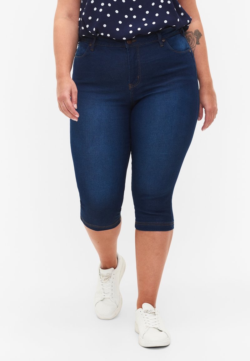 Pantalones capri de mezclilla azul oscuro con una cintura de media altura, presentando un diseño ajustado y detalles cosidos. Combinados con zapatillas blancas y una blusa de lunares.