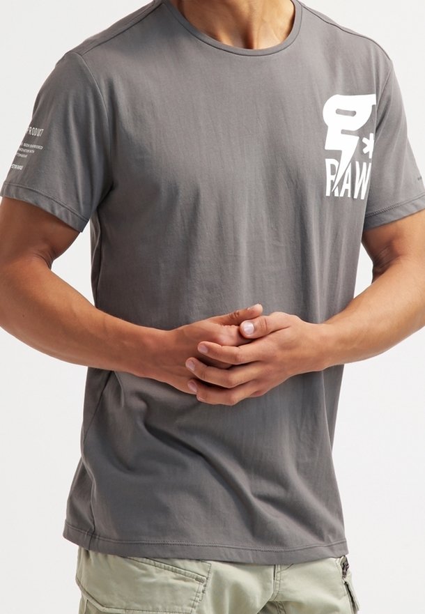 Šedé bavlněné tričko s krátkými rukávy, kulatým výstřihem, s bílým grafickým motivem na levé části hrudi a nápisem na pravém rukávu.