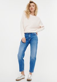 Lichtroze geribde trui met lange mouwen en een losse fit, gecombineerd met middelblauwe broek met rechte pijpen en beige sneakers met grijze accenten.
