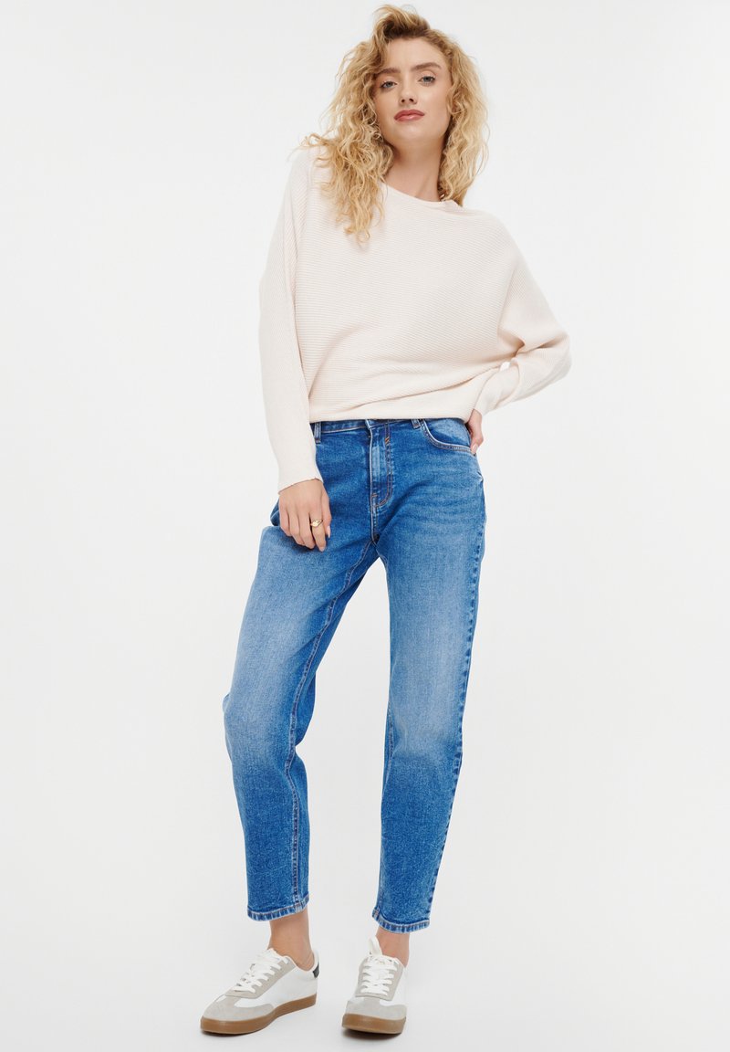 Lichtroze geribde trui met lange mouwen en een losse fit, gecombineerd met middelblauwe broek met rechte pijpen en beige sneakers met grijze accenten.