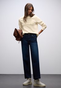 Sweatshirt crème avec le texte "Paris", jean large en denim foncé et baskets claires, tenant un sac marron contre un fond neutre.