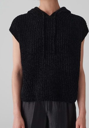 Ärmelloser schwarzer grob gestrickter Hoodie, getragen über lockeren dunklen Hosen, zeigt Oberkörper und Arme vor einfachem Hintergrund.