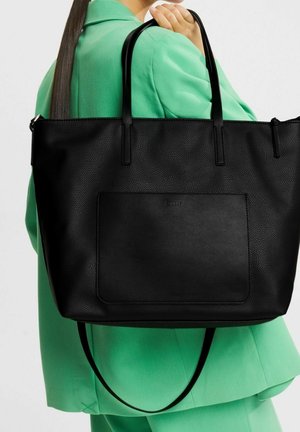 Mujer con blazer verde sosteniendo un gran bolso tote negro texturizado con bolsillo delantero y doble asa sobre el hombro.