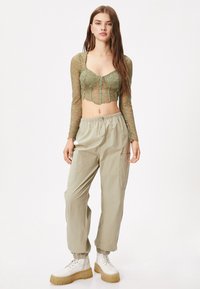 Top corto de encaje verde oliva con mangas largas y dobladillo festoneado, combinado con pantalones cargo beige con puños elásticos y bolsillos, llevados con botas contundentes blancas.