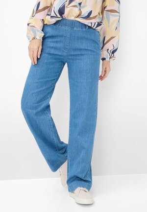 Vrouw die lichtblauwe jeans met wijde pijpen draagt, een blouse met lange mouwen en pasteltinten met patroon, en lichtgekleurde sneakers, staand met één been gebogen.
