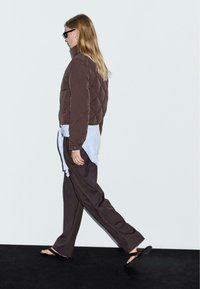Giacca trapuntata marrone, sottocamicia grigia annodata in vita, pantaloni marroni larghi e sandali neri; modello che cammina su uno sfondo neutro.