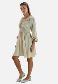 Frau steht und trägt ein beigefarbenes, langärmliges Kleid mit geraffter Taille und Knöpfen, kombiniert mit silberglänzenden, flachen Schuhen.