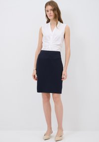 Valkoinen hihaton paita nappidetaililla, yhdistettynä tiukkaan tummansinisään kynähameeseen. Mallilla on päällään beigekutiset teräväkärkiset ballerinat ja hopeinen rannekoru.