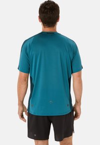 Maglietta sportiva a maniche corte in teal, con un motivo testurizzato, dettagli neri sui lati e orlo curvo. Indossata con pantaloni corti neri.