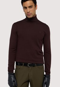 Pull en col roulé bordeaux en tissu tricoté, design ajusté, manches longues et un détail de logo subtil sur la poitrine gauche. Porté avec des gants en cuir noirs.