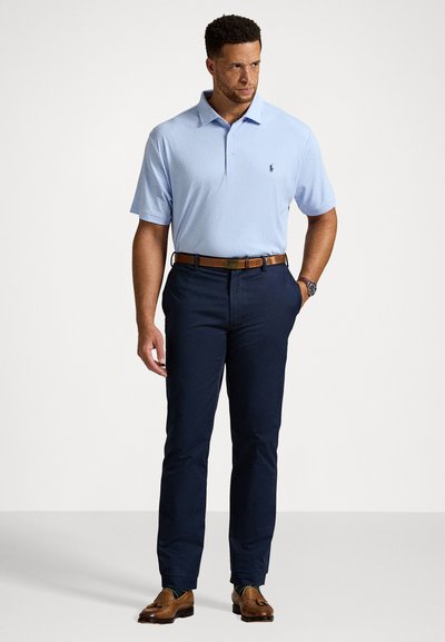 Homem com uma camisola polo azul-claro, calças azul-marinho escuras, cinto castanho, sapatos loafers castanhos com borlas, relógio, de pé com a mão no bolso contra um fundo branco.