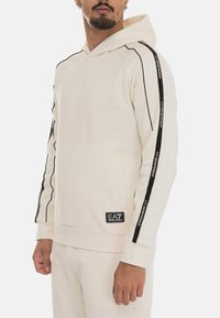 Felpa leggera beige realizzata in tessuto morbido, con dettagli a strisce nere lungo le maniche, tasca frontale e patch con logo in basso.