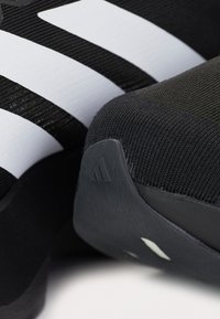 Ingrandimento di una scarpa Adidas nera con superiore in maglia, texture a coste e tre strisce bianche su sfondo di tessuto nero.