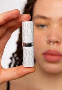 Witte lipsticktube met lichtgrijze cijfers en een zilveren band met het "Elizabeth Arden" logo, vastgehouden door een hand met op de achtergrond een vervaagd gezicht.