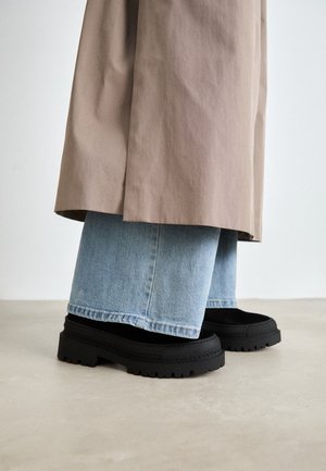 Sorte chunky-sål sko iført med lyseblå brede jeans og en beige trenchcoat, stående på et glat lyst gulv.