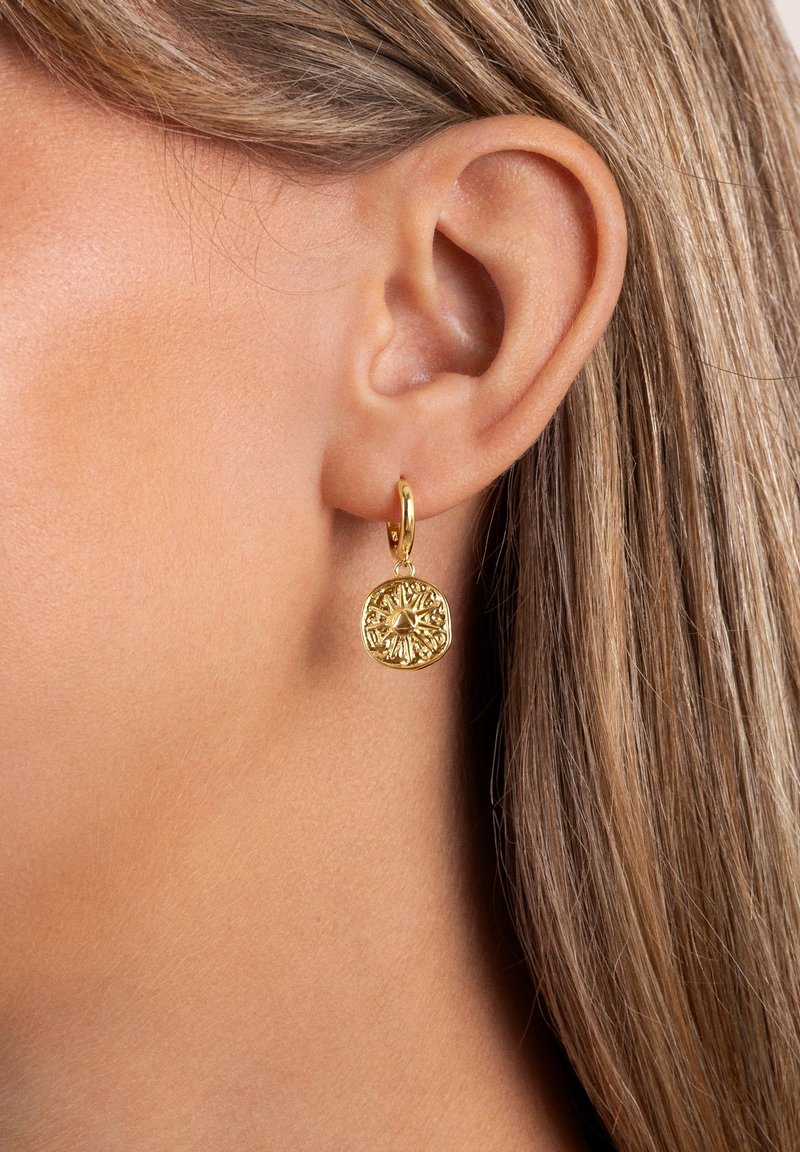 Gros plan d’une oreille portant une boucle d’oreille créole en or avec un pendentif rond texturé présentant un motif rayonnant.