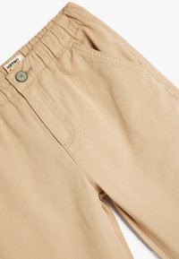 Beige lasten housut joustavalla vyötäröllä ja napin suljennalla. Ominaisuuksina etutaskut ja kevyt, sileä kangas.