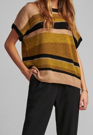 Femme portant un pull ample à rayures dans des tons marron, moutarde, noir et beige, associé à un pantalon noir, main dans la poche, et un bracelet en or.