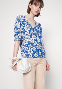 Blusa floral com fundo azul e flores brancas, design em V e mangas bufantes. Combinada com calças bejes e uma mala branca.