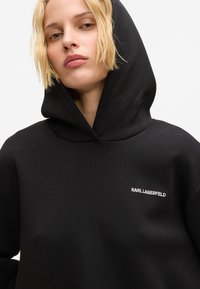 Pessoa loira a usar um hoodie preto com o logótipo "KARL LAGERFELD" no peito, olhando ligeiramente para baixo com uma expressão neutra.