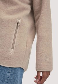 Pullover in pile beige con tasca laterale con zip, finitura testurizzata e dettaglio del logo ricamato sulla manica. Presenta polsini a coste.