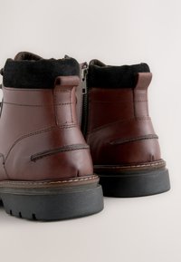 Braune Leder-Stiefeletten mit schwarzem Textilkragen, seitlichem Reißverschluss und profilloser schwarzer Gummisohle. Sauberes, minimalistisches Design.