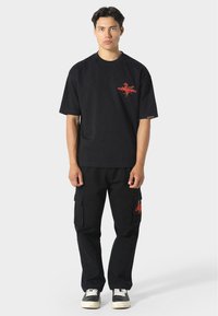 Jongeman in een zwart oversized t-shirt en cargo shorts met rode grafische ontwerpen, staande tegen een effen lichtgrijze achtergrond.