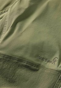 Närbild av olivgrönt tyg med texturerad sömnad och broderad "ISBJÖRN"-logotyp på friluftskläder.