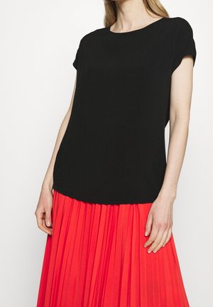 Vrouw met een zwart shirt met korte mouwen en een felrode geplooide rok, weergegeven van nek tot midden van de dij, tegen een witte achtergrond.