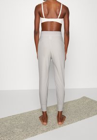 Jasnoszare, dopasowane spodnie dresowe z elastycznym paskiem i ściągaczami, wykonane z gładkiego, elastycznego materiału. Modelka stoi boso, zwrócona tyłem do aparatu.