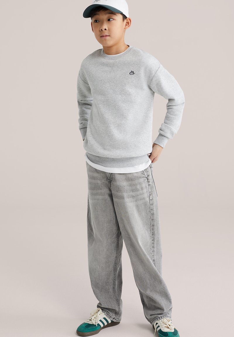 Sudadera gris con un pequeño logotipo, combinada con jeans grises holgados. Se lleva con una camiseta blanca interior y zapatillas con detalles verdes.