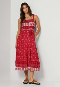 Vrouw draagt een mouwloze, rode midi-jurk met bloemige en geometrische patronen, staand met één hand in de zak en bruine sandalen.