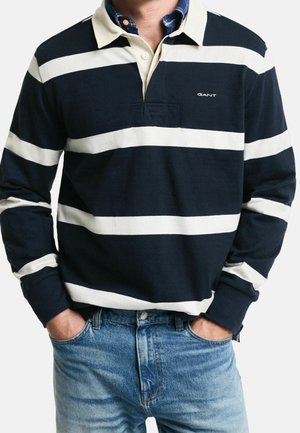 Homme portant un maillot de rugby à manches longues bleu marine avec des rayures horizontales blanches et un jean bleu, les mains dans les poches.