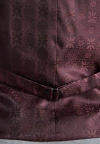 Tissu bordeaux avec des motifs floraux et circulaires intriqués, présentant une finition texturée et un fermoir noir à la taille.