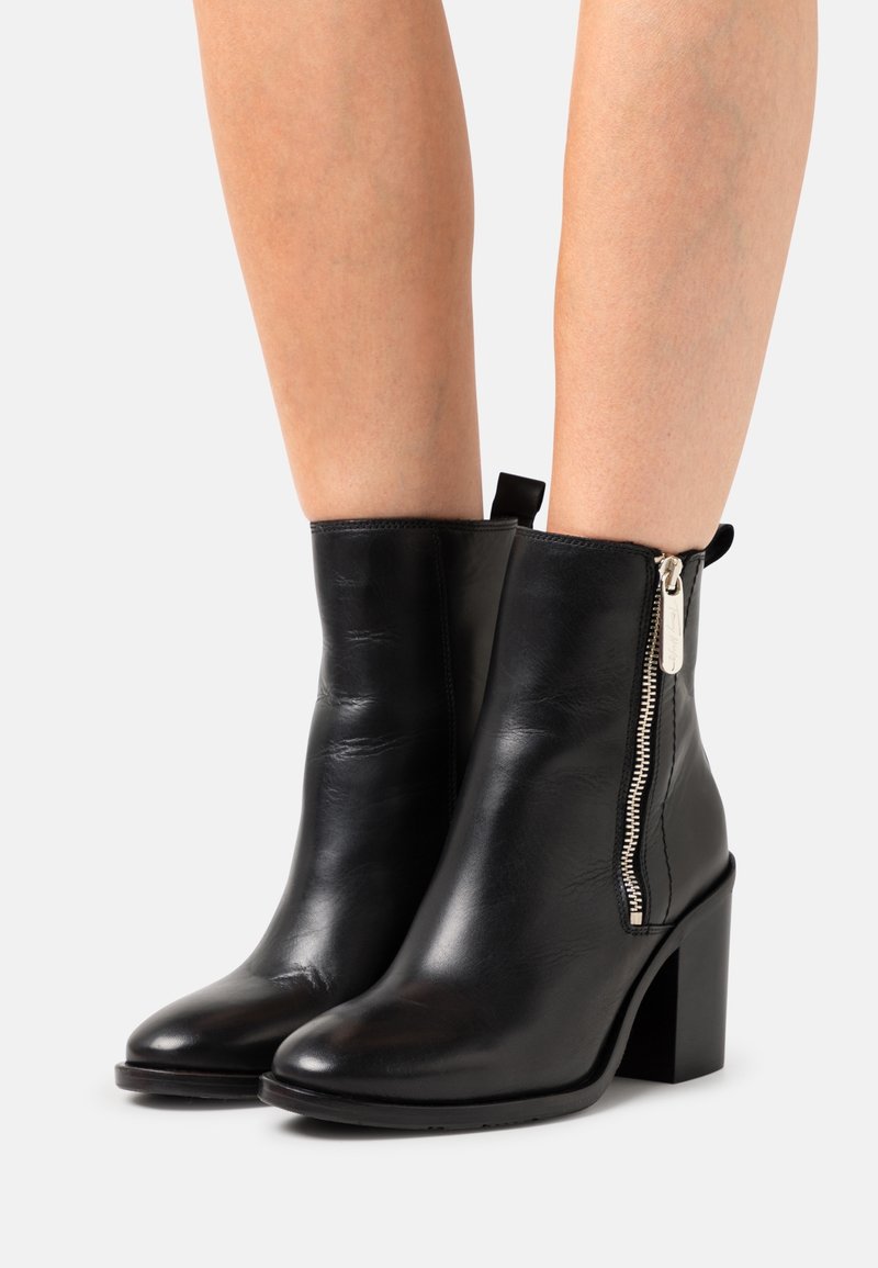 Bottines en cuir noir avec talons carrés et zips latéraux argentés portées sur des jambes nues sur un fond uni.