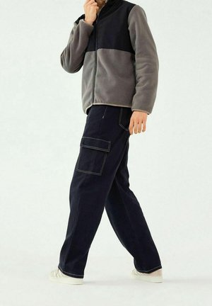 Veste polaire grise et noire avec fermeture éclair intégrale, associée à un pantalon cargo bleu marine avec plusieurs poches et coutures contrastantes. Baskets blanches.