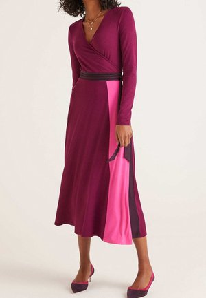 Woman wearing a long-sleeve wrap dress in dark purple with pink and black side panels, paired with matching purple and pink heels.