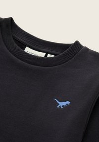 Sweatshirt noir à col rond avec un col côtelé et un petit dinosaure brodé bleu sur la poitrine. L'étiquette à l'intérieur du col indique la marque et la taille.