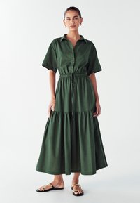 Zielona, krótko rękawowa sukienka z guzikami, z zaciśniętą talią, warstwową spódnicą i szczegółami ze sznurkiem. Połączona z czarnymi sandałami z złotymi akcentami.